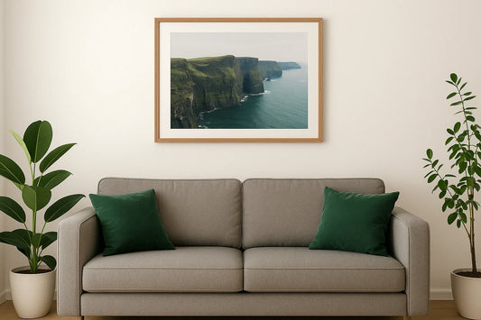 Bright Irish living room with a large framed Cliffs of Moher print centred above a grey sofa and green cushions, flanked by potted plants.