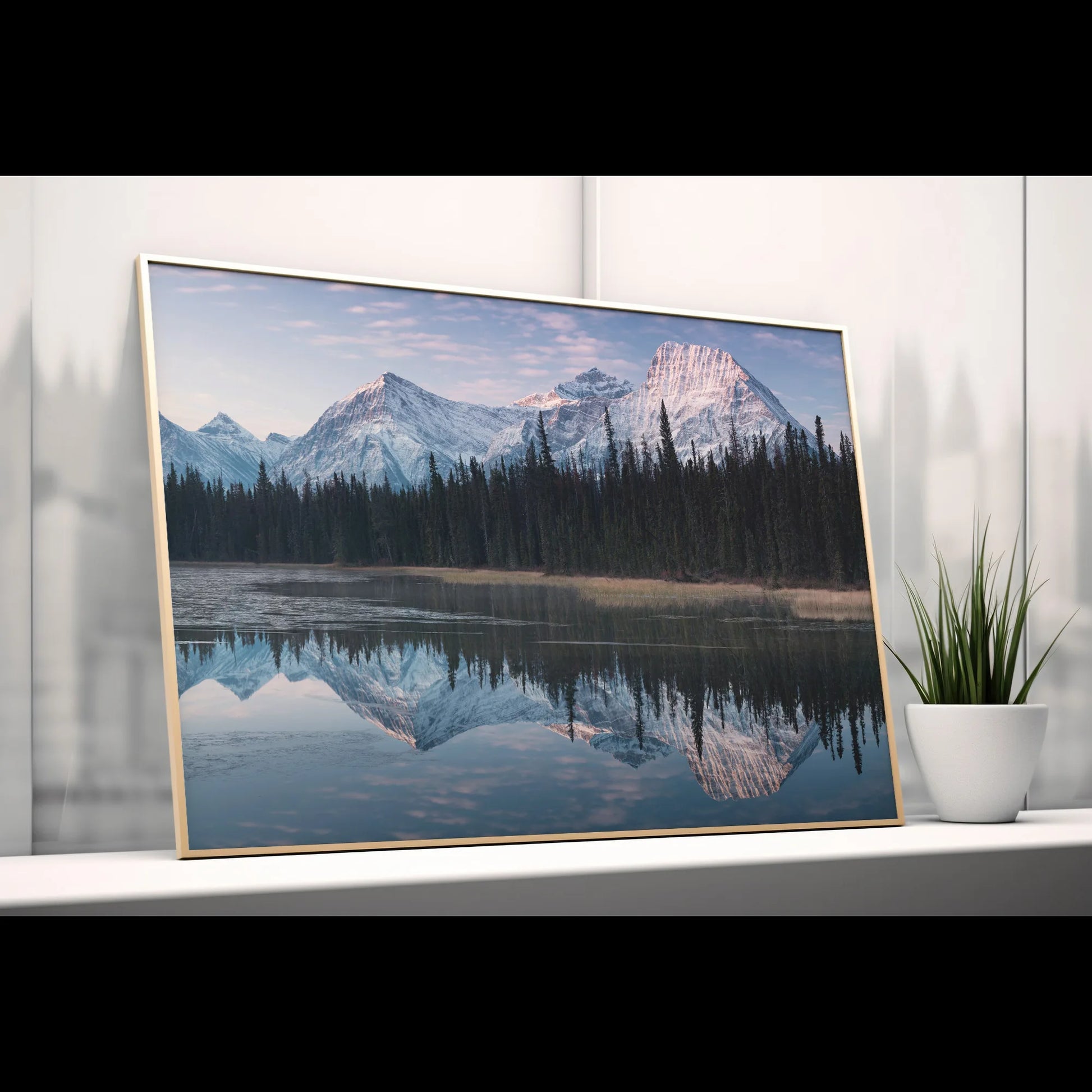 A breathtaking landscape print featuring a snow-capped mountain range reflected in a tranquil lake. The artwork captures soft pastel hues of dawn, with tall evergreens lining the shore, creating a calming and immersive scene.