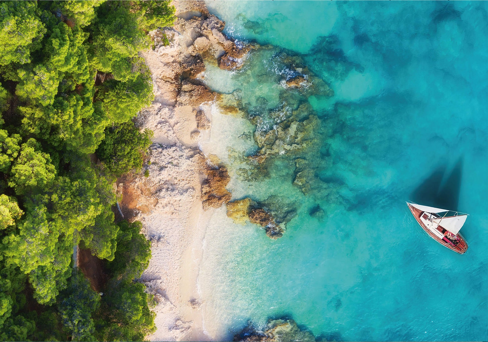 A stunning aerial view of a tropical coastline with lush green trees, golden sand, and turquoise waters. A red sailboat floats nearby.