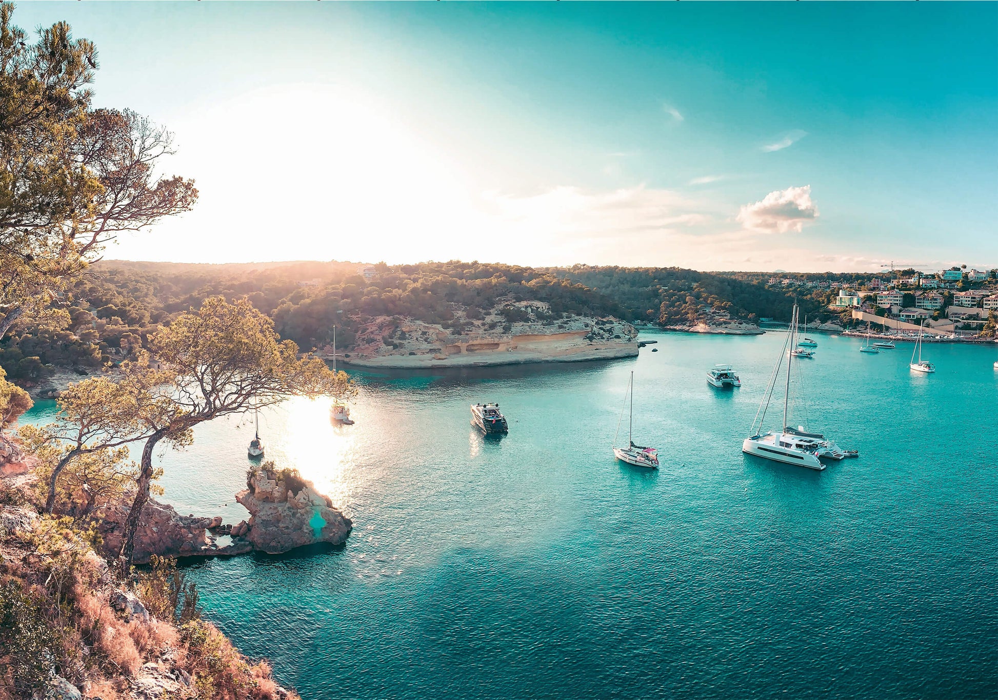 A breathtaking coastal bay scene at sunset with yachts and sailboats floating on turquoise waters. The golden sunlight casts a warm glow over the tranquil cove, surrounded by lush greenery and rocky cliffs.